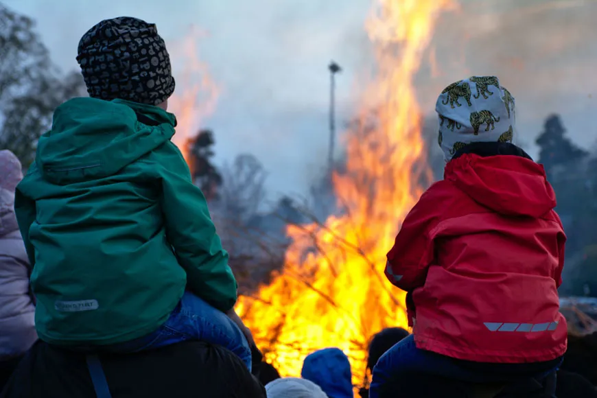 Två barn som sitter på vuxnas axlar syns bakifrån framför en valborgseld