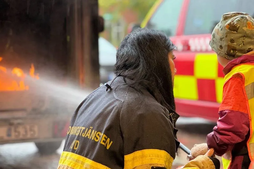 Ett barn sprutar vatten på en eld under översyn av personal från räddningstjänsten som sitter på huk bredvid barnet