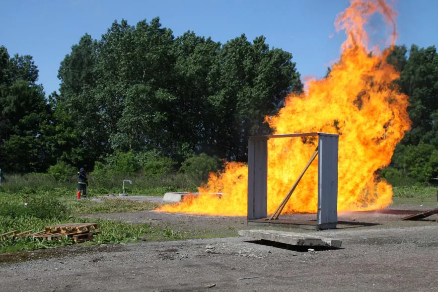 En eld brinner på en grusplan. En person i skyddskläder står i bakgrunden. 